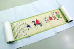 馬に乗った人とお連れの人々をカラフルな色を使って描いたイラストや文字が書かれている絵巻物を広げた写真