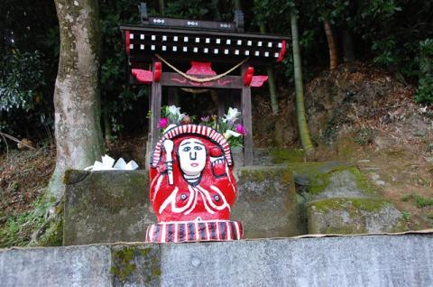 小さな鳥居の前に設置された、赤色で右側にしゃもじを握っている田の神さあの写真