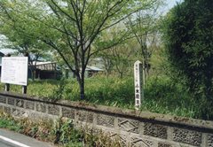 塀側の敷地内に古墳跡を示す細長い木製の案内看板が設置された、草木に覆われている小木原古墳の写真