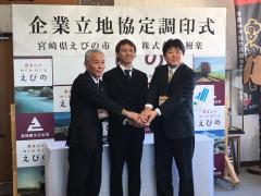 企業立地協定調印式の横断幕の下でスーツを着た男性三名が両手を重ね合っている写真