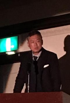 演台のマイクの前に立っているスーツを着た男性の写真