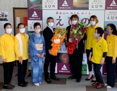 花束を持った市長と男性1名、黄色いシャツを着た女性5名、着物を着た女性1名の集合写真