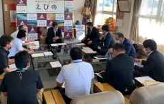 資料の置かれたガラステーブルの周りに置かれたクッション性の椅子に座っている市長と大勢の関係者の写真