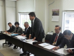 資料が置かれた長テーブルの席で市長が立って話をしており、横に関係者5名が座っている写真