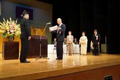 舞台上の演台前に立っている市長にスタンドマイクの前に立っている男性が感謝状を述べている写真