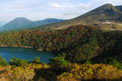 コバルトブルー色の池の周りの林がちらほら紅葉しており、遠くに山並みが見えている写真