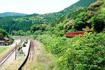 駅前の線路と駅の上の山並みの線路を走る赤い列車の写真