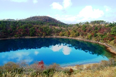 池の水がコバルトブルー色をしている不動池の周りに木々が生い茂っており、後方に韓国岳が見えている写真