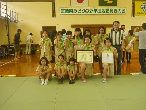 緑色のスカーフを付け探検服を着た子ども達と関係者の男性が賞状と景品を持ち前列の子どもは体育座り、後列の子ども達は立ってピースサインをして笑顔で写っている集合写真