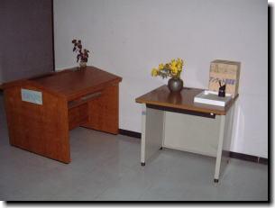左側に茶色の台、その右横に花瓶に生けられた花と箱が置かれた机がある傍聴手続き所の写真