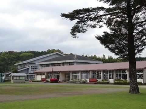白い雲と森を背景に、灰色、黒、赤の3つの大きな屋根から成る校舎を、芝生と松の木があるグラウンド越しに撮影した写真