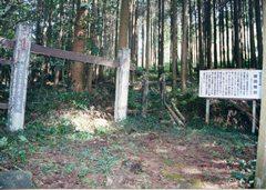 林の中の右側に説明看板が設置され、左側に門の様な柵が設置されている関跡の写真