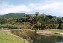 奥の山並みと手前の川との間に小高い丘がある飯野城跡の写真