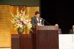 金屏風が置かれた舞台上でスーツ姿のえびの市長が挨拶をしている写真