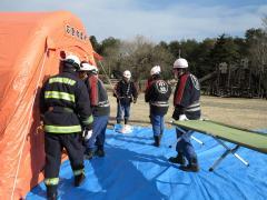 青色のビニールシートの上にオレンジ色のテントが設置されており、防災訓練をしている5名の消防団員の写真