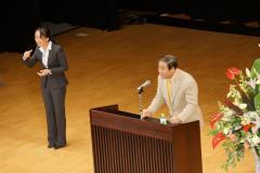 ステージの演台前で話をしている男性と、左端に立って手話をしているスーツを着た女性の写真