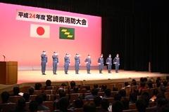 背景のスクリーンがピンクの舞台上で制服を着た消防団員達が敬礼をしている写真