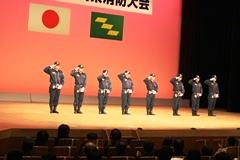 舞台の奥に日本の国旗、宮崎県旗が掲げられていて、8名の消防団員が敬礼をしている写真