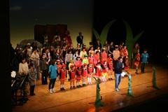 舞台上に子供や大人の出演者たちが並んでいて、前方に代表の男性が立って挨拶をしている写真