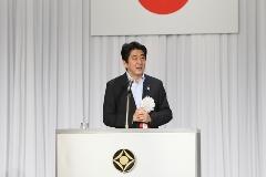 安部首相が白い幕に掲げられた日本国旗の前で挨拶をしている写真