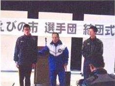中央に青いジャージを着た年配の男性、両脇に黒いジャージを着た男性二人が立っている写真
