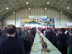 日本国旗が掲げられた舞台が奥にあり、参加者達が立って舞台を見ている様子を後ろ側から写している写真