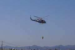 空の中央に一台のヘリコプターが飛んでいて、ヘリコプターから人が吊るされている防災訓練の写真