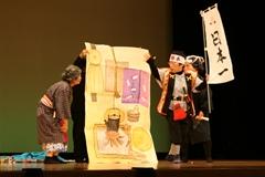 舞台上で日本一と書かれたのぼり旗を持ち桃太郎の恰好をした人や着物を着たおばあさんの恰好をした人が演劇をしている写真