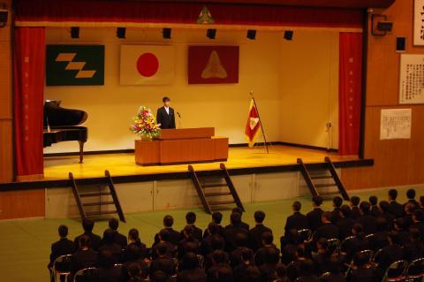 体育館ステージの演台前に立ち話をしている男性とステージ下で椅子に座っている生徒達を後方から写した写真