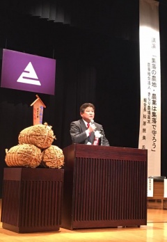 えびの市章が飾ってあるステージ上で米俵の隣の演台前でマイクを持ち、話をしている市長の写真