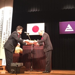 日本国旗とえびの市章が飾ってあるステージ上で市長が白髪の男性に品物を渡している写真