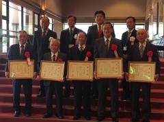 赤いじゅうたんの階段で額縁に入った賞状をもって立っている5名の男性とその後段に立っている市長と3名の男性の集合写真
