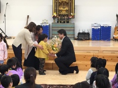 ステージ上で黄色いシャツを着た子供と二人の女性が市長に花束を手渡している写真