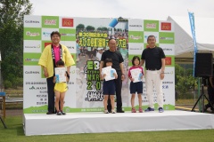 芝生の上に設置されたステージ上に立っている3名の子供と2名の男性、市長の写真