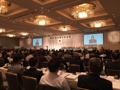 会場前方に掲げられている日本国旗の両端にあるプロジェクタスクリーンを見ている大勢の参加者を後方から写した写真