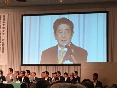 プロジェクタスクリーンに元内閣総理大臣の安倍晋三さんが映し出され、その下に座っている大勢の参加者の写真