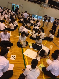 体育館でグループに分かれ、白い模造紙を広げて文字を書いている大勢の学生の写真