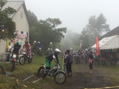 霧の中をバイクに載っている大勢のライダーと白いテントの周辺から見ている観客の写真