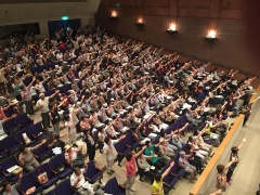 会場内で観客席に座り、手を挙げている大勢の人々を上から写した写真
