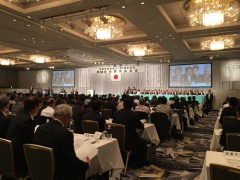 ステージ上に日本国旗と、スクリーンが掲げられており、前方を見て、椅子に座っている大勢の参加者を会場後方から写した写真