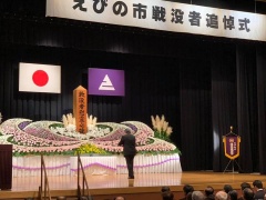 えびの市戦没者追悼式と書かれた横断幕の掲げられたステージにきれいな花祭壇が設置され、スーツ姿の男性が祭壇の前に立っている写真