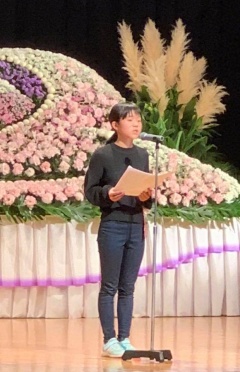 ステージ上に設置された花祭壇の前で黒い洋服を着た女の子が作文を披露してる写真