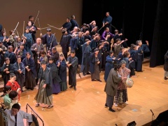 ステージ前方で舞台衣装の着物を着て提灯を持っている男女と後方では袴を着て棒を持って武芸をしている大勢の演者が写っている公演の様子の写真