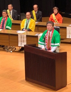 ステージ後ろに設置された長机に座る緑色や黄色の法被を着た男性や黄色のタスキを肩に掛けている男女、講演台の前に立って話をしている緑の法被を着た市長の写真