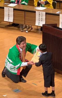 緑の法被を着た市長が片膝をつき、黒いフォーマルスーツを着た男の子に笑顔で品物を手渡している写真