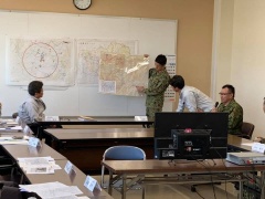 ホワイトボード前で大きな地図を持っている自衛官とマイクをもって座って説明している自衛官、地図を見ながら熱心に説明を聞いている職員の方々の写真