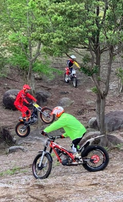 大きな岩や木の生えている林の中の斜面を、バイクに乗った3人のライダーが走っている写真