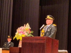 綺麗な檀上花が飾られている講演台で、消防団の制服を着ている市長が話をしてる写真