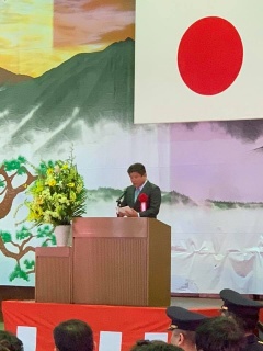 日本国旗の飾られたステージの講演台に立ってスピーチをしている市長の写真