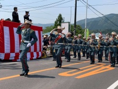 紅白の幕のステージ上に立つ人に向かって敬礼しながら行進しているえびの駐屯地の音楽隊のパレードの写真